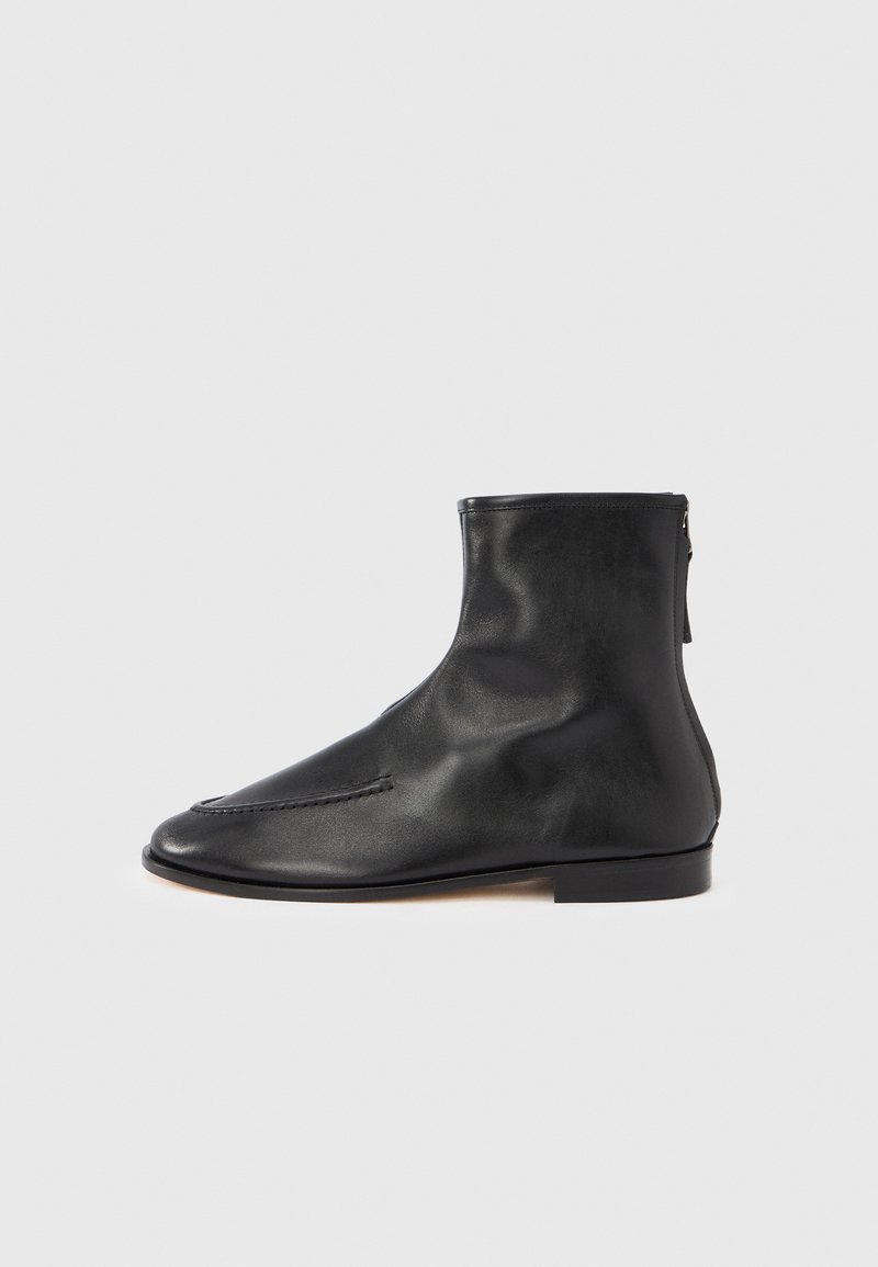 Stivaletto alla caviglia in pelle nera con finitura liscia, punta appuntita, dettagli di cuciture minime e piccola cerniera posteriore per la chiusura. Tacco piatto.