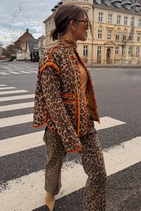Luipaardprint outfit in bruin met oranje accenten; omvat een jacket met contrasterende afwerking en bijpassende broek. Gesloten beige laarzen.