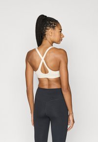 Bēša sporta krūšturis ar krustveida aizmugures dizainu, platām siksnām un izgriezuma detaļu. Melnas augsta vidukļa legingi papildina apģērbu.