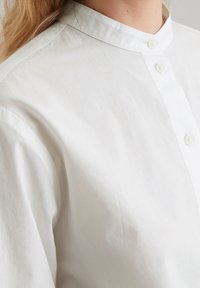 Valkoinen napillinen paita mandariinikauluksella, jossa on sileä pinta ja minimalistinen muotoilu. Kaksi etunappia näkyvissä.