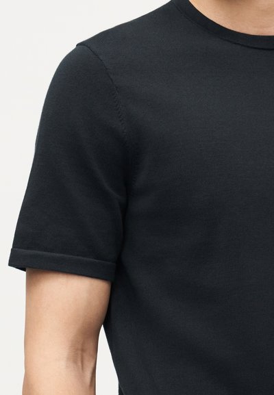 Άνδρας που φοράει εφαρμοστό, κοντομάνικο, σκούρο ναυτικό μπλε πλεκτό μπλουζάκι με στρογγυλή λαιμόκοψη, μπροστά σε απλό φωτεινό φόντο.