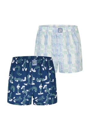 Muskelöser Mann mit kurzen geflochtenen Haaren und Bart, der dunkelblaue Boxershorts mit weißen und grünen abstrakten Mustern trägt und die Hände hinter dem Rücken hält.