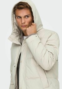 Beige pufferjack met een grote capuchon, ritssluiting en drukknoopsluiting, en twee zijzakken. Het materiaal lijkt glad met een gestikte textuur.