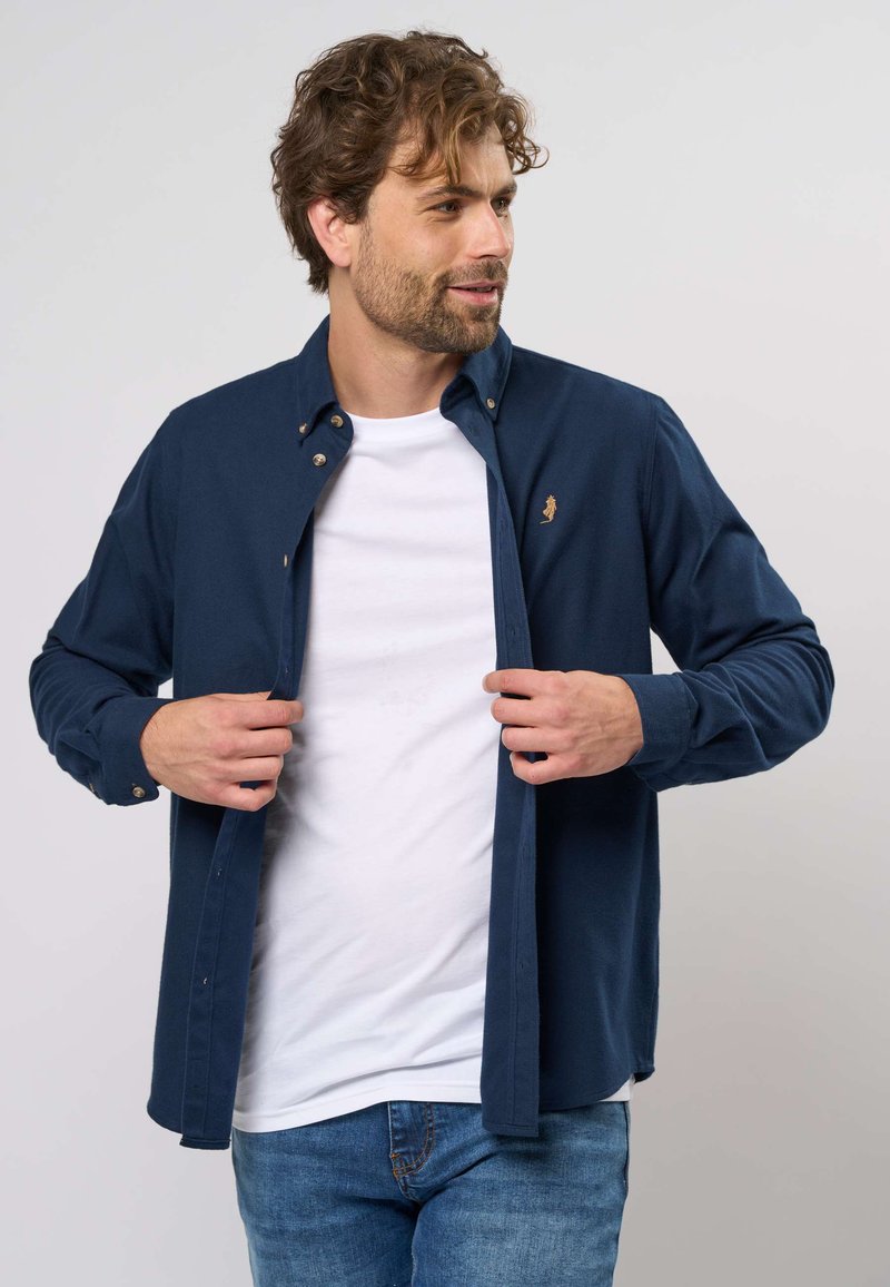 Tamsiai mėlyna mygtukinė marškiniai iš minkštos medžiagos, su subtiliu logotipu krūtinėje ir ilgomis rankovėmis, uždėta ant baltos marškinėlių.