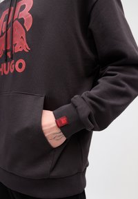 Mørkegrå hettegenser med et rødt grafisk design, kengurulomme, ribbestrikkede mansjetter og et logo-patch på ermet. Mykt, strukturert stoff.