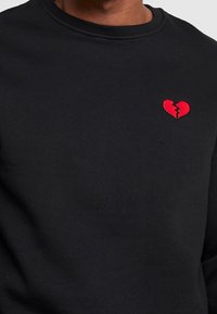 Czarna bluza z bawełny z małym, czerwonym, haftowanym logo złamanego serca na lewej piersi. Standardowy krój z okrągłym dekoltem.
