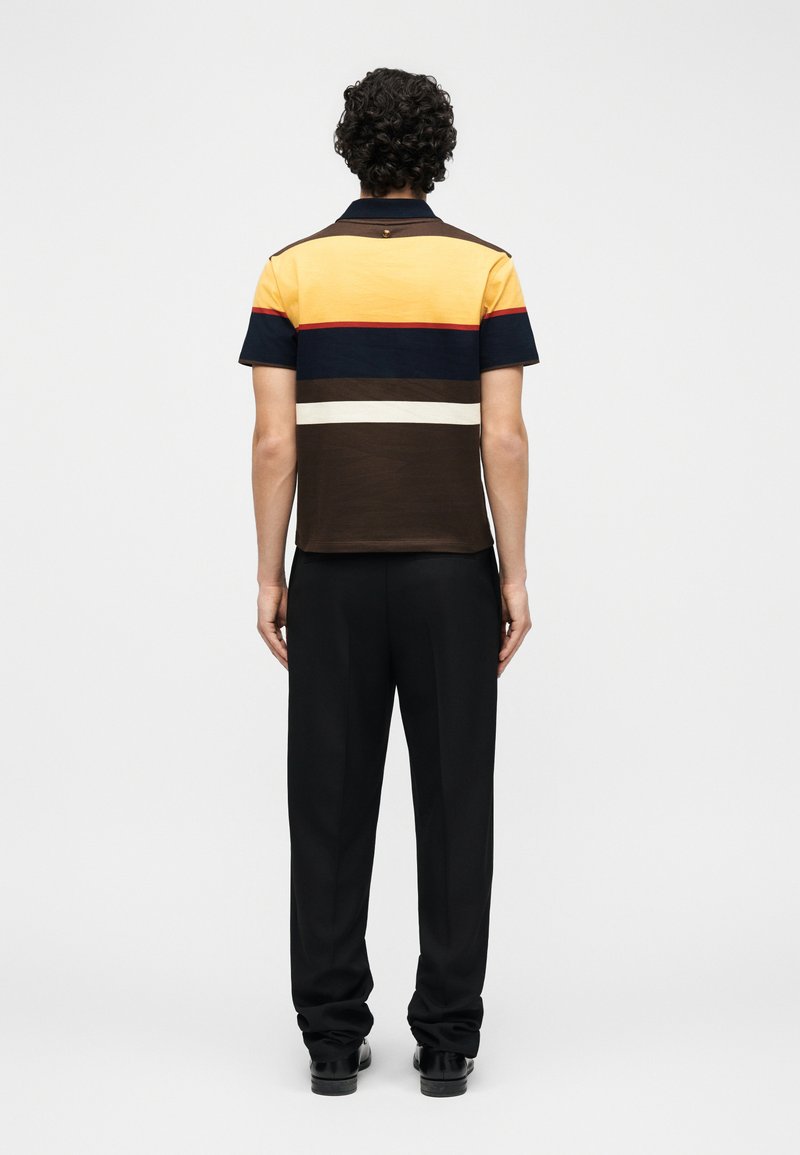 Man with curly hair wearing a short-sleeve polo shirt with yellow, navy, brown, white stripes and black pants, facing away on white background.