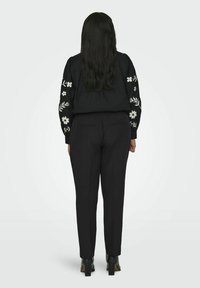 Melnā izšūtā jaka ar ziedu motīviem uz piedurknēm, kopā ar melniem šauriem biksēm un potītēm, skatījums no aizmugures.