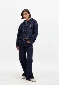 Denims jaka ar kapuci, tumši zila, saīsināts dizains, lielas priekšējās kabatas, sudraba pogas, kopā ar atbilstošām platās kājas biksēm, baltas kurpes.