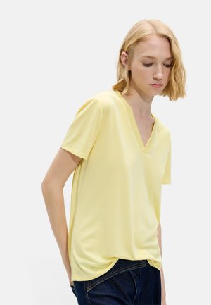 Młoda osoba o blond włosach do ramion, ubrana w jasnożółtą koszulkę z krótkim rękawem i dekoltem w serek oraz ciemnoniebieskie dżinsy, patrząca w dół.