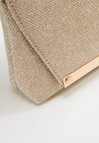 Gros plan sur un coin de pochette texturée en paillettes dorées avec un accent métallique doré brillant, posée sur une surface blanche.