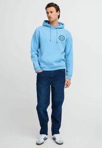 Felpa azzurra con un grafiche di un sorriso sul petto, abbinata a pantaloni di jeans blu scuro e sneaker bianche con dettagli neri.