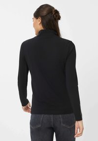 Frau mit dunklen Haaren in einem Dutt trägt ein schwarzes Langarmoberteil und dunkle Jeans, mit dem Rücken zur Kamera vor einem schlichten weißen Hintergrund.