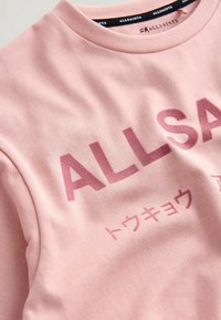 Vaaleanpunainen puuvillaneule, jossa on suuret "ALLSAINTS"-kirjaimet. Suunnittelussa on hienovaraista tekstuuria ja lisätekstiä japanilaisilla merkeillä.
