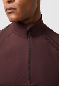 Bordeauxfarbene sportliche Zip-Up-Jacke aus glattem, dehnbarem Stoff. Mit hohem Kragen und subtil auf der Seite aufgedrucktem BOGNER-Logo.