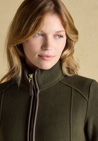 Woman with blonde hair wearing an olive green fleece jacket with a zip and high collar, looking slightly to the side.