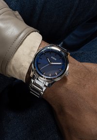 Montre en acier inoxydable avec un cadran texturé bleu, des accents argentés et un bracelet à maillons. Portée sur un poignet avec une manche claire.