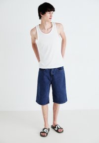 Débardeur blanc, shorts en jean bleu et claquettes vertes avec des lettres blanches. Le mannequin est debout, les mains dans les poches, sur un fond neutre.