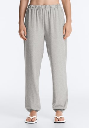 Personne portant un pantalon de jogging gris clair à taille élastique avec des poignets élastiques et des sandales blanches, debout devant un fond blanc uni.