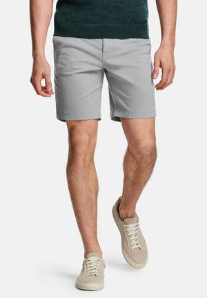 Homme mannequin portant un short gris clair, un haut vert et des baskets beiges à lacets, marchant devant un fond blanc uni.