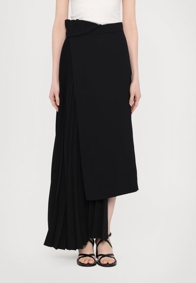 Gonna maxi plissettata nera con design drappeggiato, dotata di una vita elastica arricciata e uno spacco laterale, abbinata a sandali neri con cinturino.