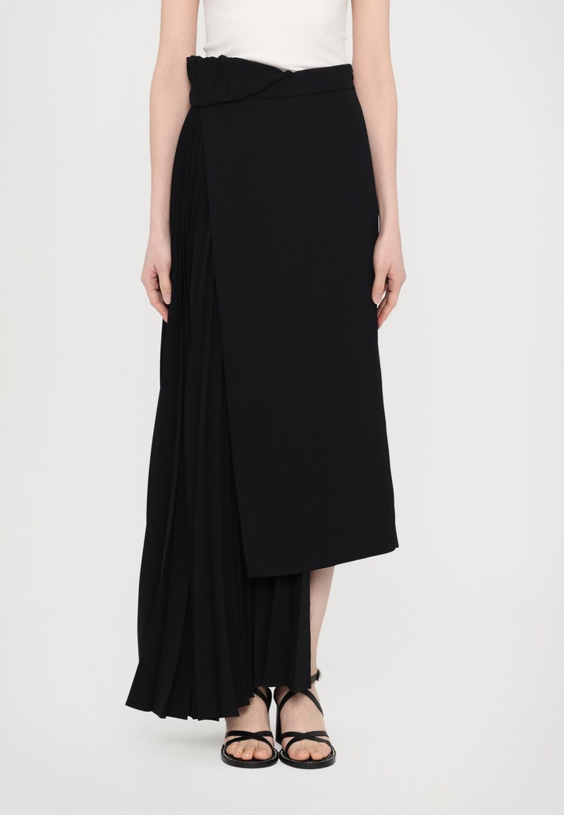 Gonna maxi plissettata nera con design drappeggiato, dotata di una vita elastica arricciata e uno spacco laterale, abbinata a sandali neri con cinturino.