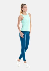 Kobieta stojąca, ubrana w jasnoniebieską bluzkę bez rękawów, ciemnoniebieskie legginsy i białe trampki, uśmiechająca się i patrząca w bok.