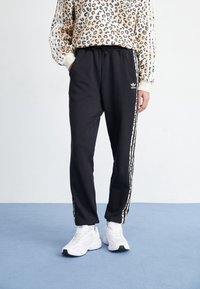 Pantaloni da allenamento neri con cintura elastica, con strisce laterali bianche e logo adidas. Abbinati a una maglietta beige con stampa leopardata.