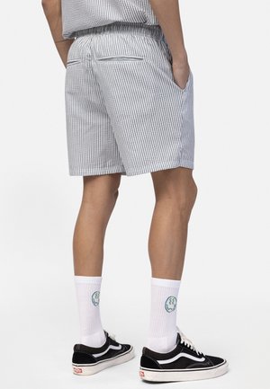 Homme portant un short rayé gris et blanc, des chaussettes blanches avec un logo visage souriant, et des baskets noires, debout les mains dans les poches.