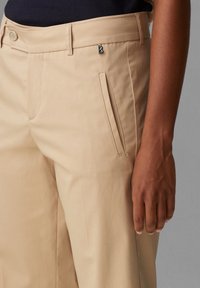 Pantaloni beige con tessuto liscio, design su misura e tasca laterale. Un'emblema silver è attaccato vicino alla vita.