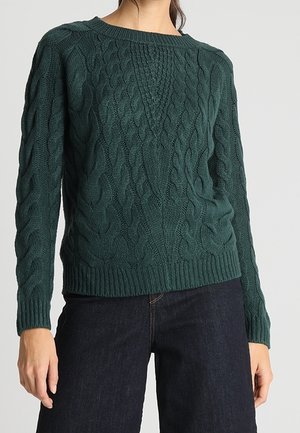 Femme portant un pull en maille torsadée vert foncé et un jean bleu foncé, debout devant un fond clair uni.