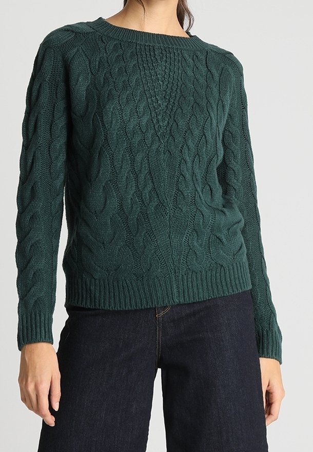 Femme portant un pull en maille torsadée vert foncé et un jean bleu foncé, debout devant un fond clair uni.