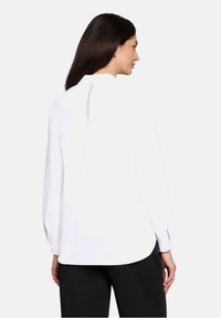 Blouse blanche à manches longues avec un col rond, présentant un détail subtil dans le dos et une coupe décontractée, associée à un pantalon noir.