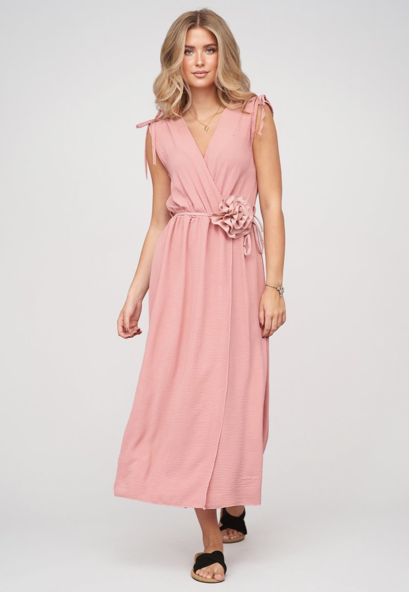 Femme blonde portant une robe portefeuille sans manches rose poudré avec un détail fleur sur le côté, debout sur un fond neutre avec des sandales noires.