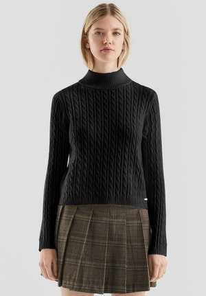 Jonge vrouw die een zwarte kabelgebreide coltrui en een bruin geruite geplooide rok draagt, staand tegen een effen achtergrond.
