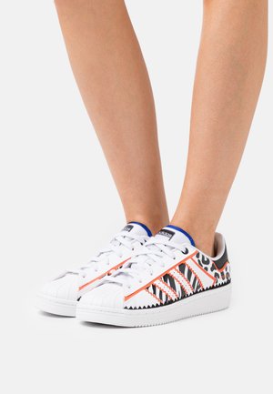 Persona con zapatillas Adidas blancas con patrones en negro, naranja y estampado de leopardo, de pie sobre una superficie blanca.