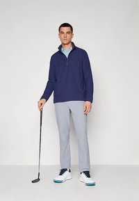 Homem seguro com um putter de golfe, vestindo um pulôver azul-marinho de fecho de zíper, uma polo azul-clara, calças cinza-claro e sapatos de golfe brancos.