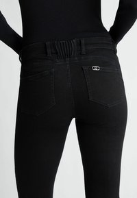 Gros plan d'une personne portant un jean noir taille haute avec deux poches arrière, dont une avec un petit détail de logo argenté.