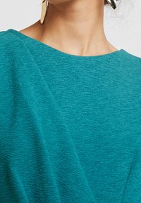 Sinisävyiset teksturoidut kangasverhot rennolla istuvuudella ja leveällä pääntiellä. Kiinnitettyinä metalliset korvakorut, jotka heijastavat valoa.