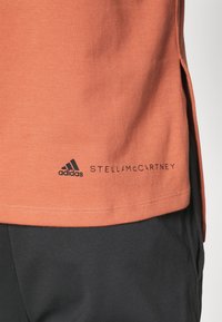 Kortärmad, orange t-shirt i bomull med rundad sidsöm. Har svarta logotyper för "adidas" och "STELLAMCCARTNEY" nära botten. Slät textur.