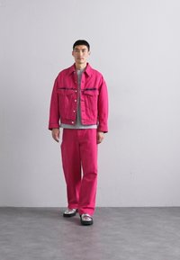 Φωτεινό ροζ τζάκετ από τζιν με δύο εμπρόσθιες τσέπες, σε συνδυασμό με παντελόνι του ίδιου χρώματος. Το μοντέλο φοράει μια γκρίζα μπλούζα από κάτω. Ασημένια παπούτσια με πλατφόρμα.