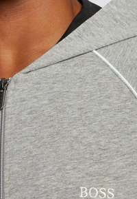 Sudadera con capucha gris con cremallera, de textura suave, con una costura de acento blanca y el logo "BOSS" bordado en blanco en la parte inferior del frente.