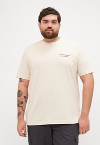 Beige bomull t-skjorte med korte ermer, rund hals, og et lite trykket logo på venstre bryst. Glatt tekstur, avslappet passform.