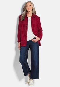 Rood en zwart blokkerig overhemdjack, witte t-shirt eronder, blauwe wijde jeans en witte sneakers; lang haar, handen op de heupen.