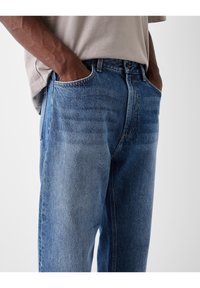 Jeans in denim blu con vestibilità rilassata, cuciture visibili e tasche laterali. Il tessuto presenta una texture scolorita con una finitura di lavaggio medio.