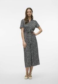 Robe florale noire avec des manches courtes, un devant boutonné, une taille cintrée, de longueur mi-haute et un design fluide. Portée avec des chaussures de couleur crème.