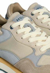Zapatillas de ante beige y gris con cordones crema, que cuentan con un panel de malla y el logo "HOF" en la lengüeta. Suela de goma con detalles en marrón claro.