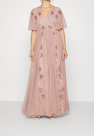 Kvinde iført en gulvlængde, støvet lyserød kjole med gennemsigtige flagermusærmer og blomsterapplikationer med palietter ned foran.