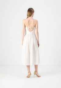 Vestido midi blanco sin mangas con un corsé ajustado, cintura fruncida y tirantes cruzados en la espalda. Presenta detalles de lazo y botones.
