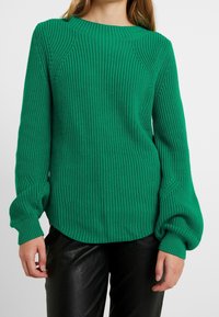 Zelený pletený svetr s žebrovanou texturou, kulatým výstřihem a volným střihem. Má balónové rukávy a mírně zakřivený lem.